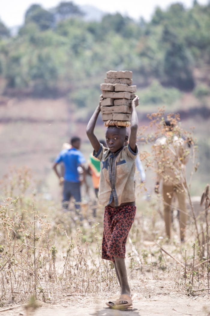 creative African child carries stack of bricks on head in rural area, underlining themes of labor and resilience.